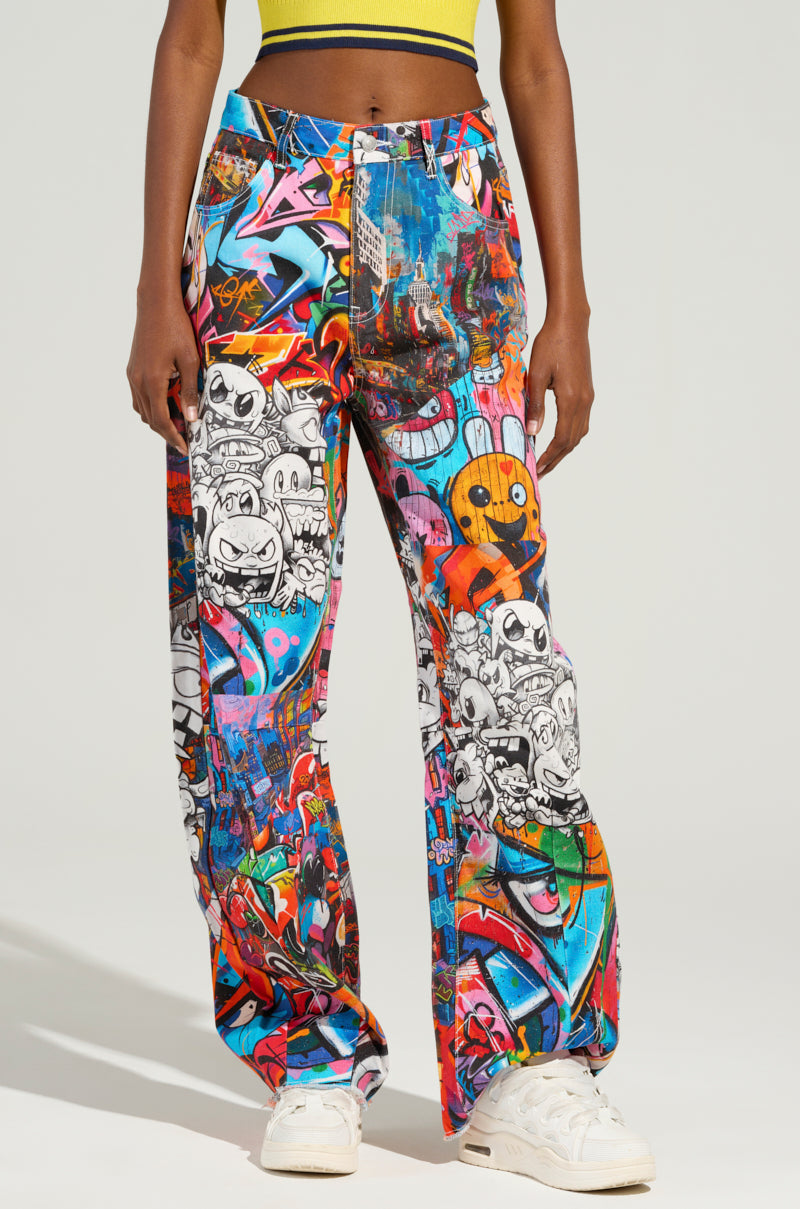 In front of a neutral background, a person dons the SAMMIE CARTOON DENIM JEANS, featuring graffiti-inspired prints paired with white sneakers. The vibrant designs showcase animated characters and abstract art, adding a playful touch that evokes street artistry to the outfit.