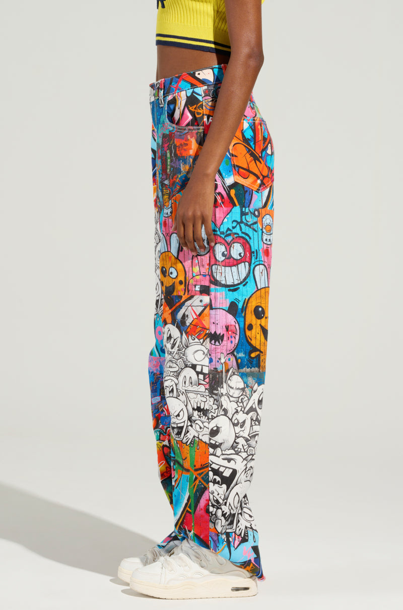 A person wearing the SAMMIE CARTOON DENIM JEANS, which showcase a vibrantly colorful, graffiti-inspired print with various cartoon and abstract designs, stands sideways. They pair these high-rise wide-leg jeans with a yellow cropped top and white sneakers.