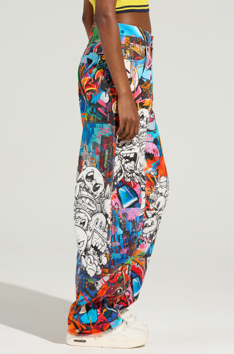 The woman is styled in the SAMMIE CARTOON DENIM JEANS, featuring a graffiti-inspired design with vibrant red, blue, and black elements set against a light backdrop. She pairs them with a yellow top and white sneakers while standing sideways.