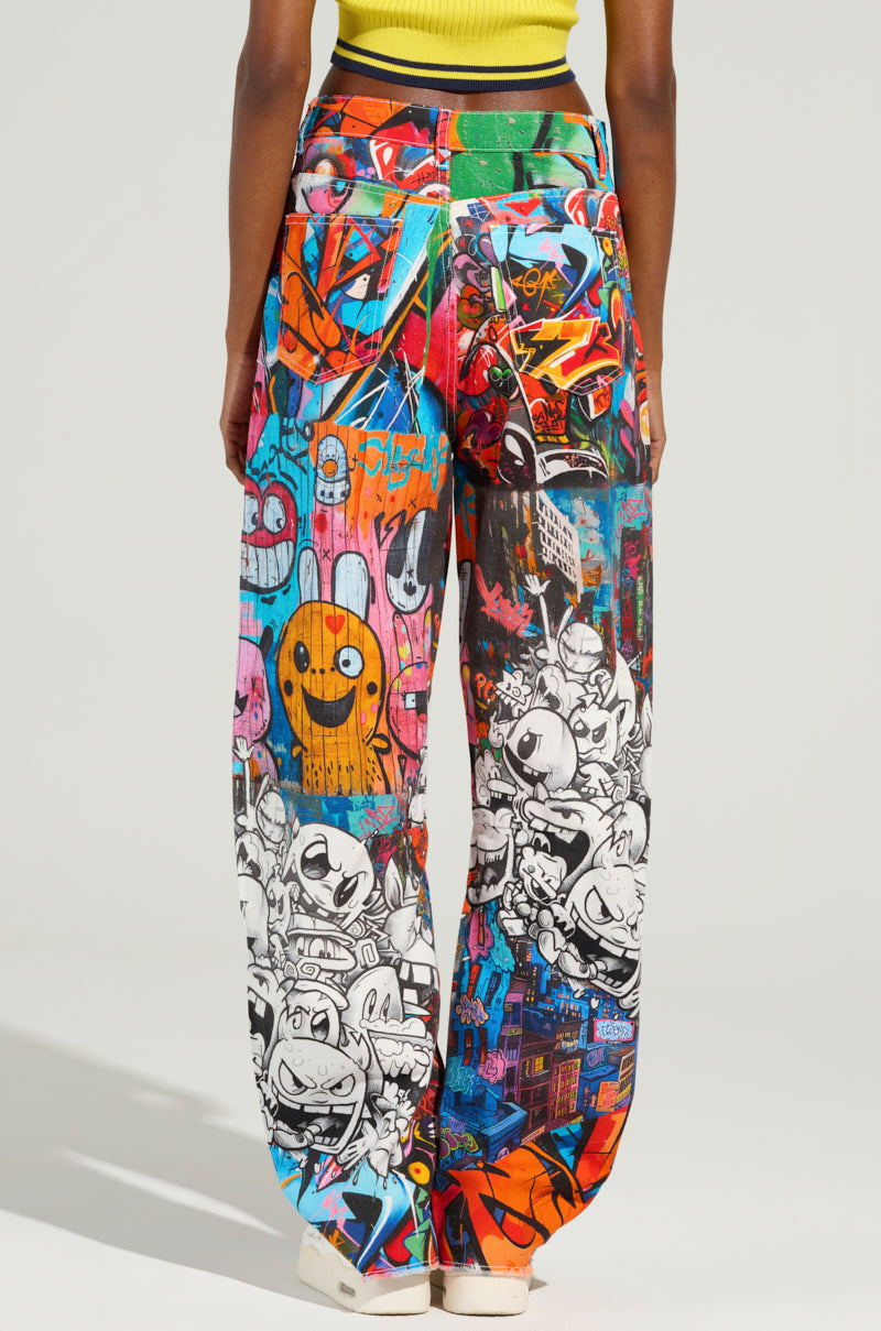 A person wearing the SAMMIE CARTOON DENIM JEANS, which are graffiti-inspired with cartoon characters and abstract designs. These high-rise wide-leg jeans showcase bright colors and whimsical patterns such as smiley faces and artistic elements. They're styled with a yellow top and white shoes.