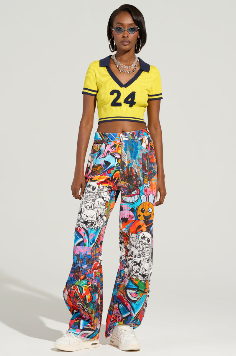A person wearing a yellow crop top with "24" on it, paired with SAMMIE CARTOON DENIM JEANS, sunglasses, and necklaces stands against a plain background. They are also wearing white sneakers.
