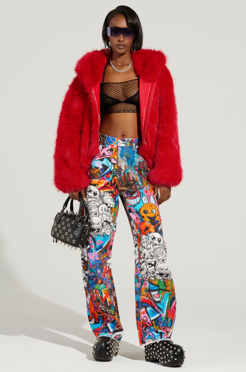 A person stands confidently, wearing a red fur jacket and black mesh top, paired with the vibrant SAMMIE CARTOON DENIM JEANS. They accessorize with sunglasses, a spiked handbag, and studded shoes, combining edgy and colorful fashion elements.