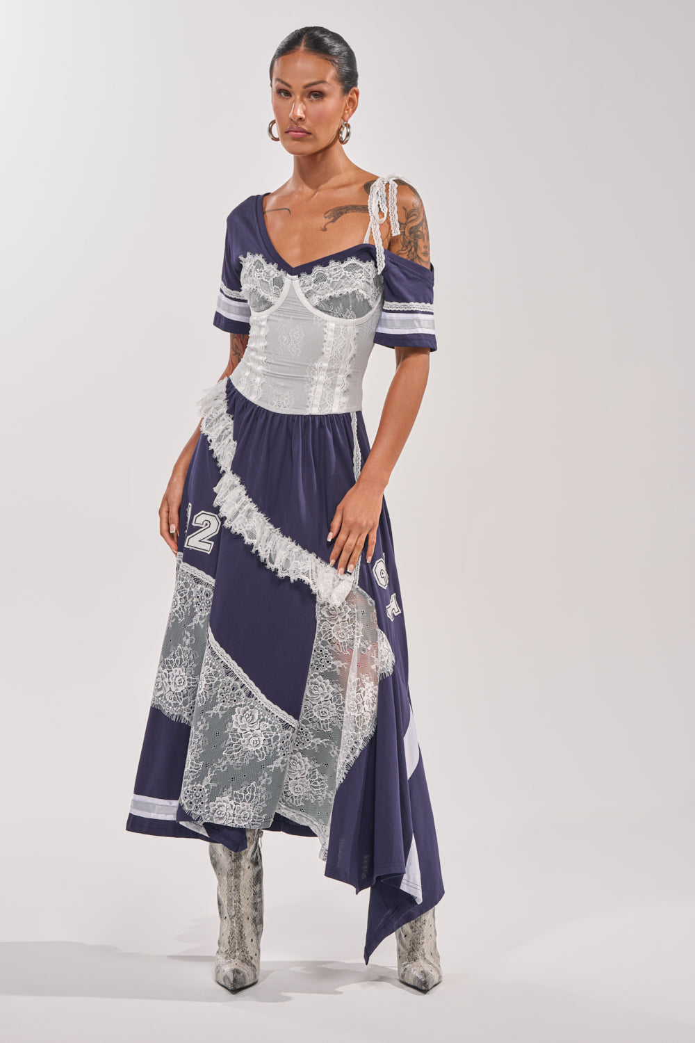 A woman poses against a white background in the BOX SUITE LACE JERSEY MAXI DRESS, a navy varsity-style dress with white lace accents and an asymmetrical hem, styled with hoop earrings, tattoos, and white snakeskin boots. 1