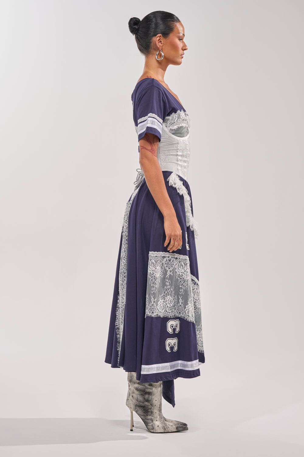 A woman in profile wears the BOX SUITE LACE JERSEY MAXI DRESS—a navy varsity-style dress with white lace—paired with a structured corset, silver high-heeled boots, hoop earrings, and her hair in a bun against a plain light background. 3