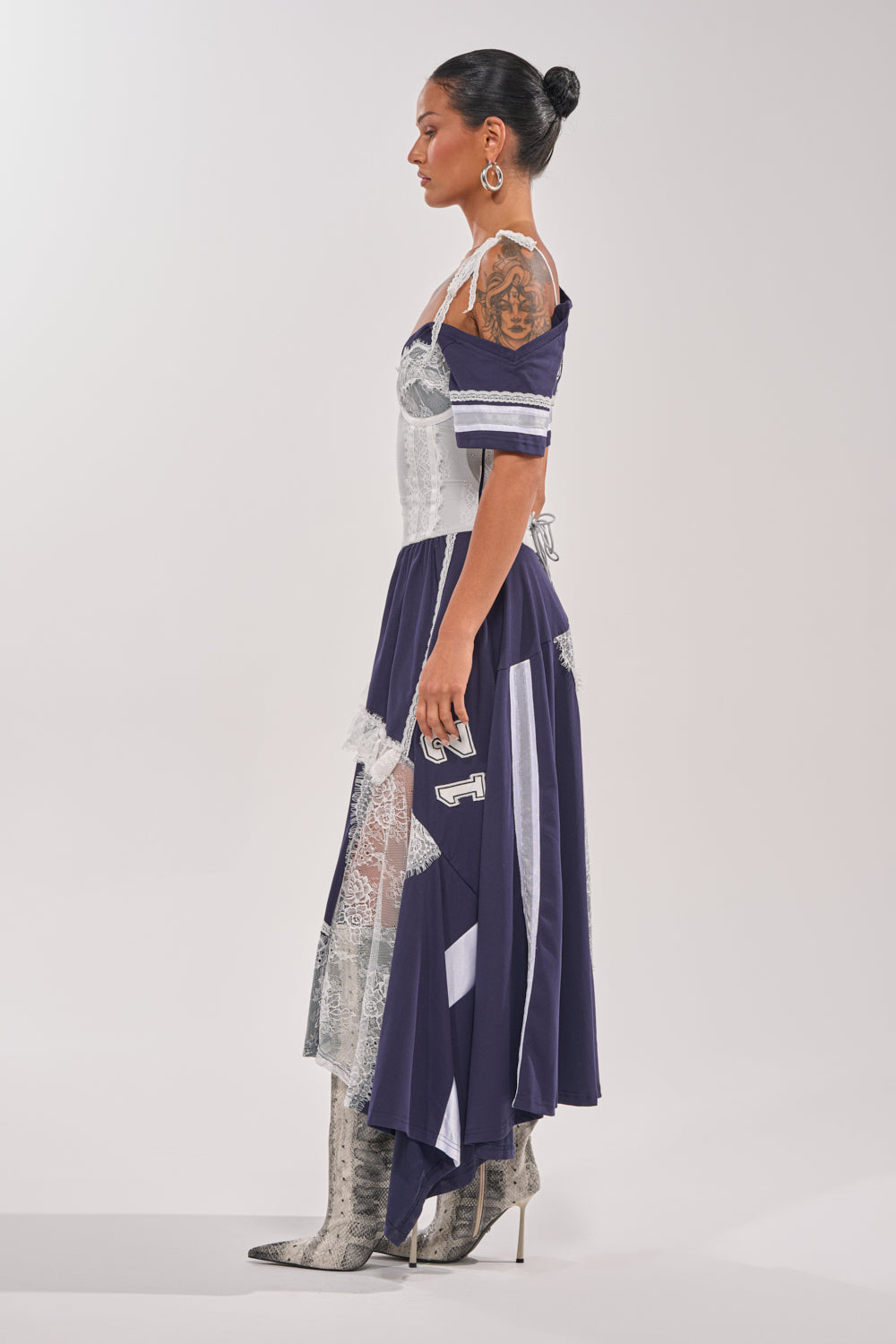 A woman stands in profile wearing the BOX SUITE LACE JERSEY MAXI DRESS, a varsity-style navy and white asymmetrical dress with lace accents and "24" on the skirt, paired with knee-high snakeskin boots and large hoop earrings. 4