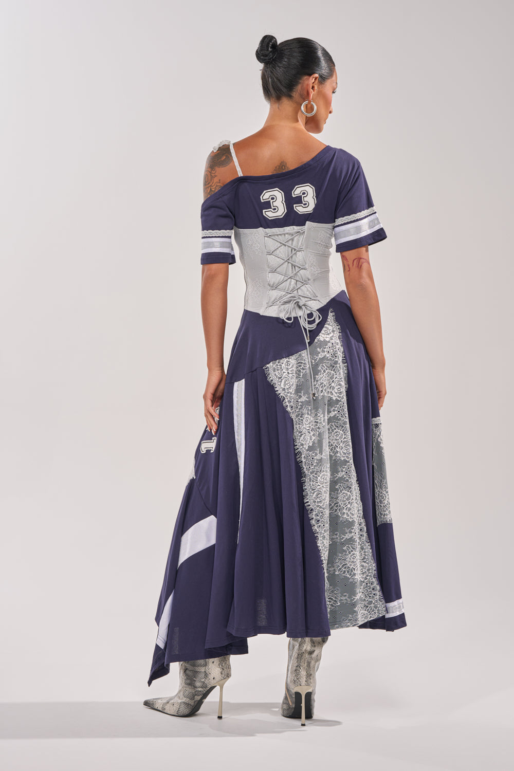 A woman faces away, wearing the BOX SUITE LACE JERSEY MAXI DRESS in navy blue with white accents and number 33 on the back. She pairs it with a gray corset, snakeskin boots, and has her dark hair styled in a bun. 5