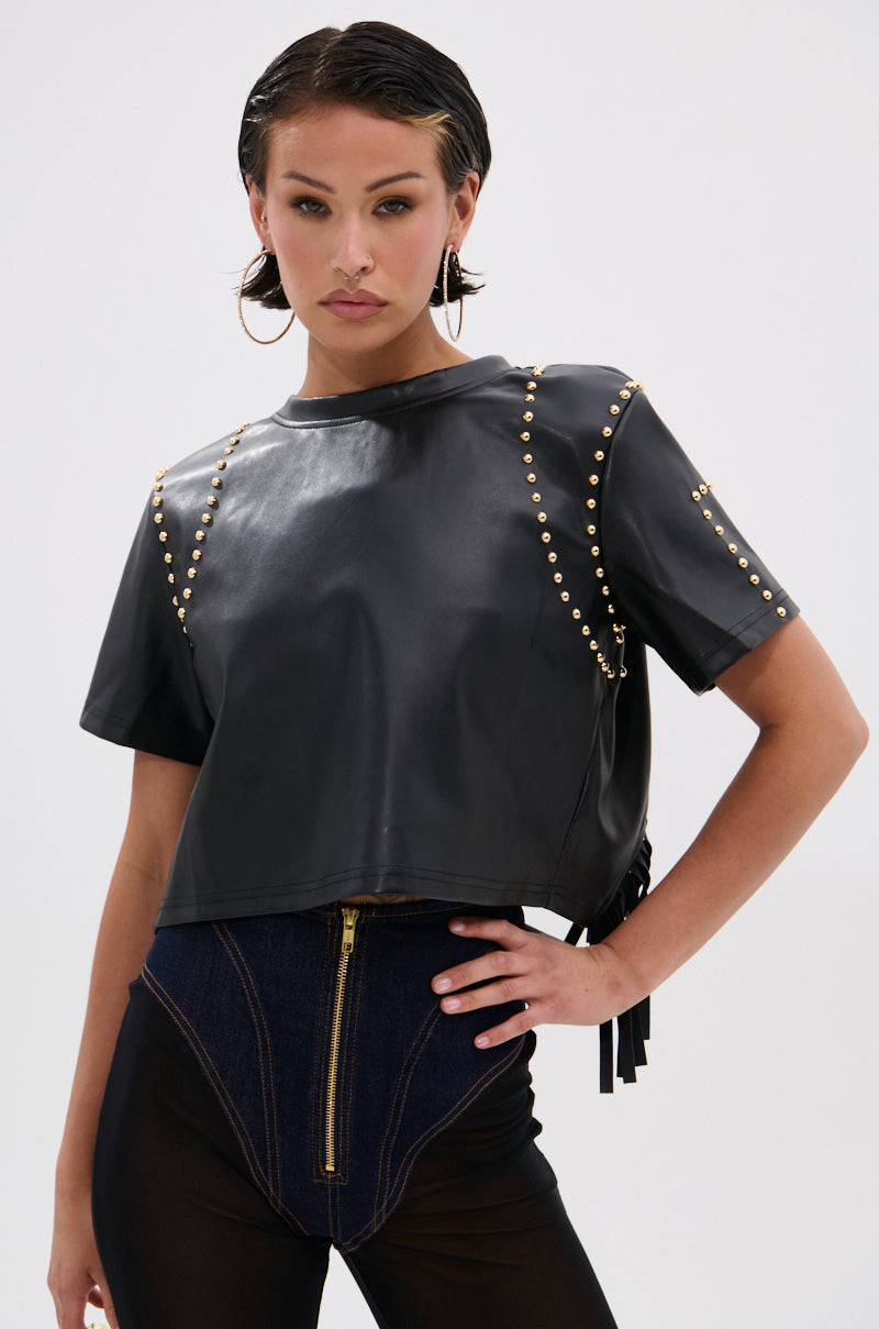 A person poses confidently in the ROCKSTAR STUDDED FRINGE TOP, a black faux leather crop with gold studs, paired with high-waisted black pants featuring a front zipper and denim accents. Slicked-back hair and hoop earrings complete the bold look. 0