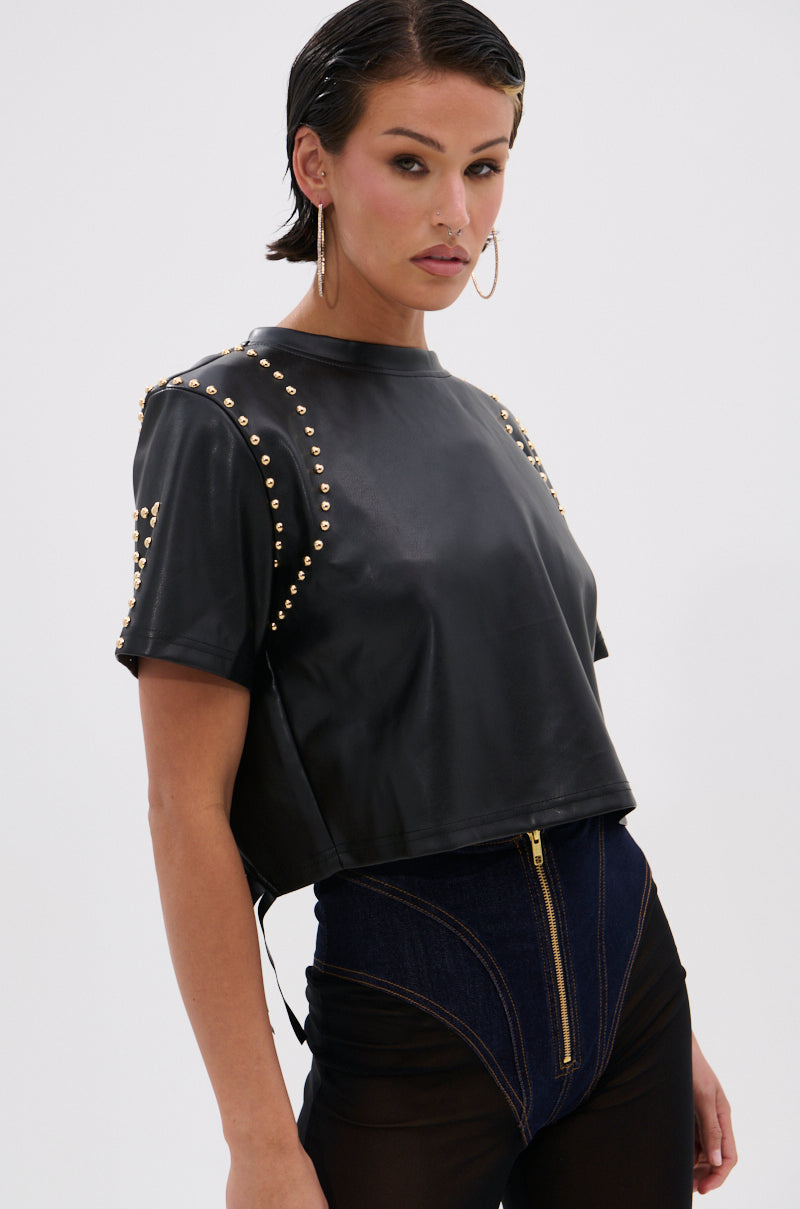 A person wearing the black ROCKSTAR STUDDED FRINGE TOP with gold shoulder and sleeve studs, paired with high-waisted dark denim pants featuring a front zipper, stands against a plain white background. 1