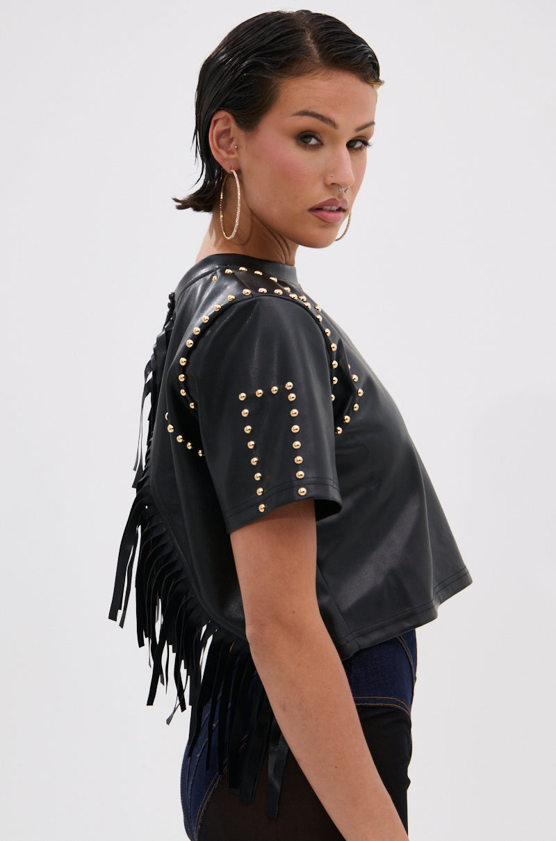 A person with short dark hair and large hoop earrings models the ROCKSTAR STUDDED FRINGE TOP, featuring black faux leather and gold studs, against a plain light background. 2