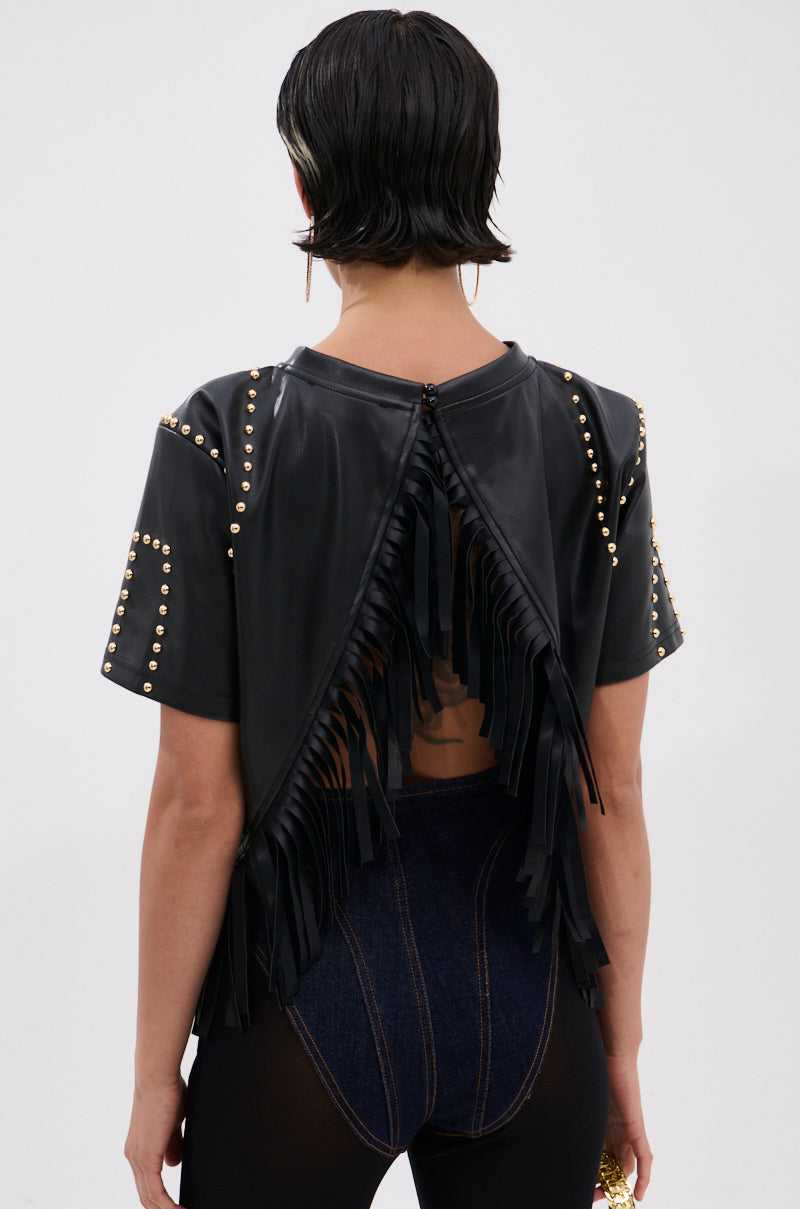 A person with short dark hair is seen from behind, wearing the ROCKSTAR STUDDED FRINGE TOP featuring black faux leather, gold studs, fringe details, and a back slit that reveals part of the lower back. Paired with dark high-waisted pants. 3
