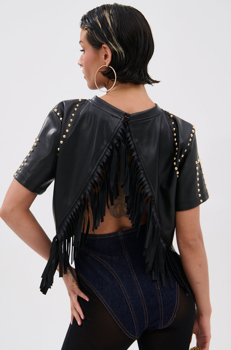 A person with slicked-back hair wears the ROCKSTAR STUDDED FRINGE TOP—a black faux leather crop top with gold studs and fringe—paired with high-waisted dark denim. The open back reveals a bold tattoo. 4