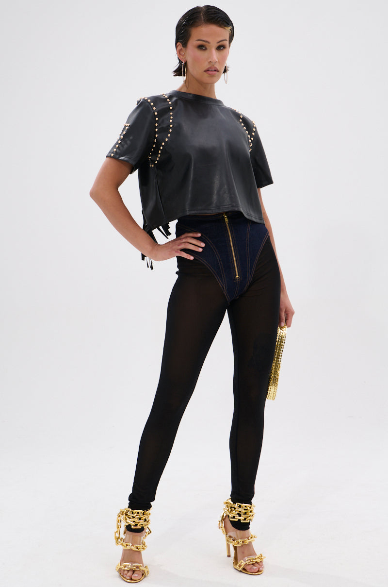 A woman poses confidently in the ROCKSTAR STUDDED FRINGE TOP, paired with high-waisted dark blue shorts, sheer black leggings, gold chain heels, and a small gold bag against a plain white background. 6