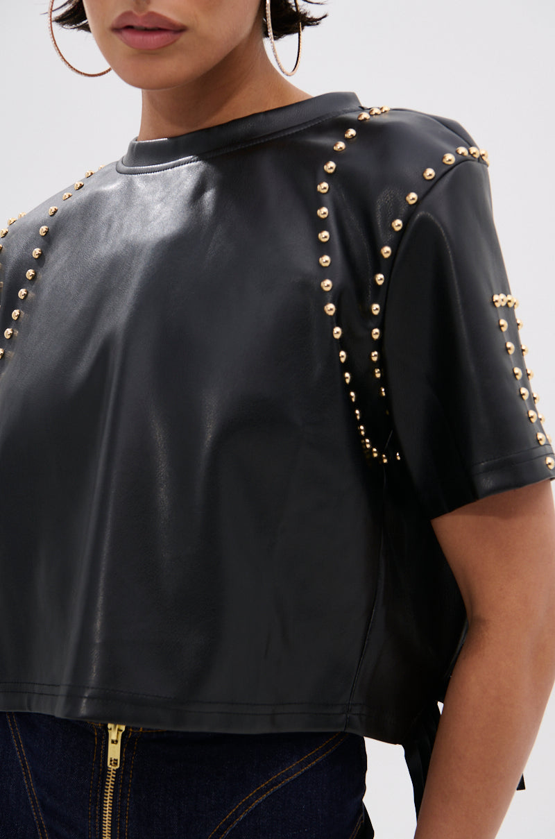 A woman wears the ROCKSTAR STUDDED FRINGE TOP— a black faux leather, short-sleeve crop top with gold studs on the shoulders and sleeves—paired with dark denim jeans featuring a visible gold zipper. 7