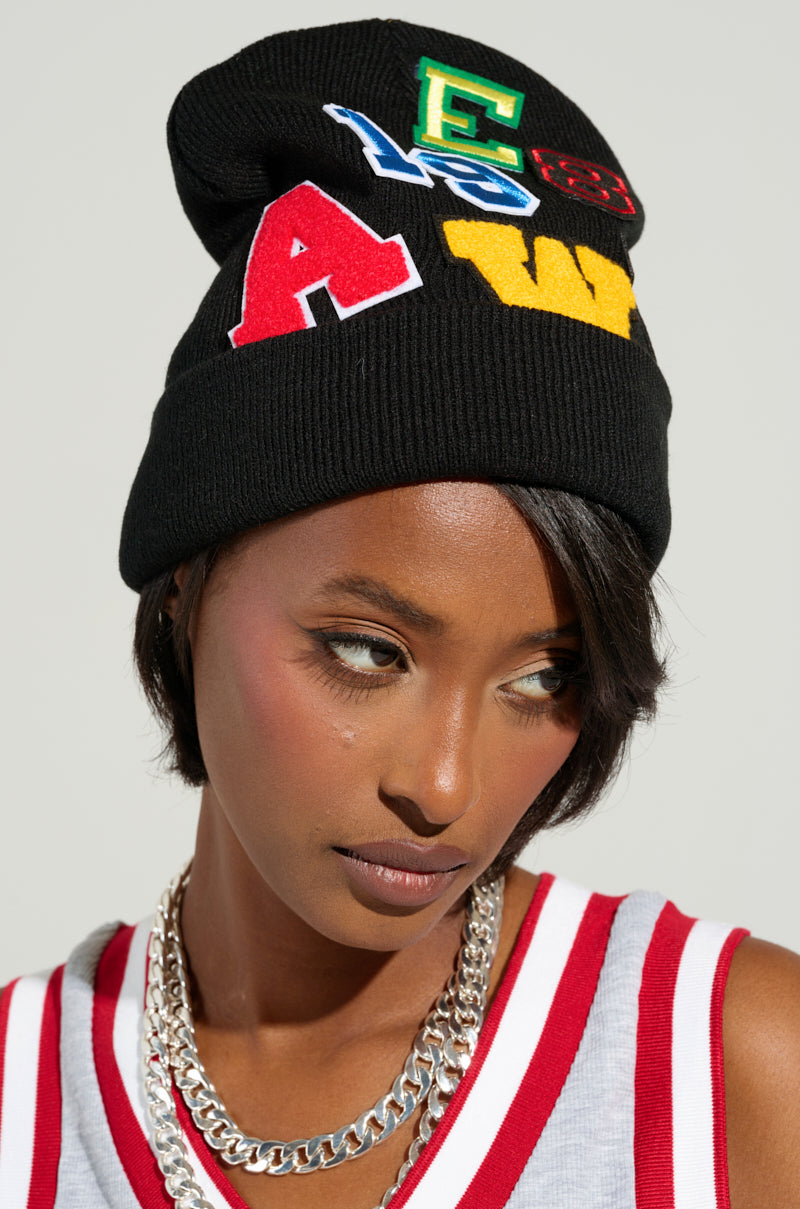 A person wearing the SPORTS VIBES PATCH BEANIE, which features letterman embroidered patches, sports a serious expression. They pair it with a sleeveless red and white striped top to evoke strong sports vibes and complete the look with a chunky silver chain necklace.