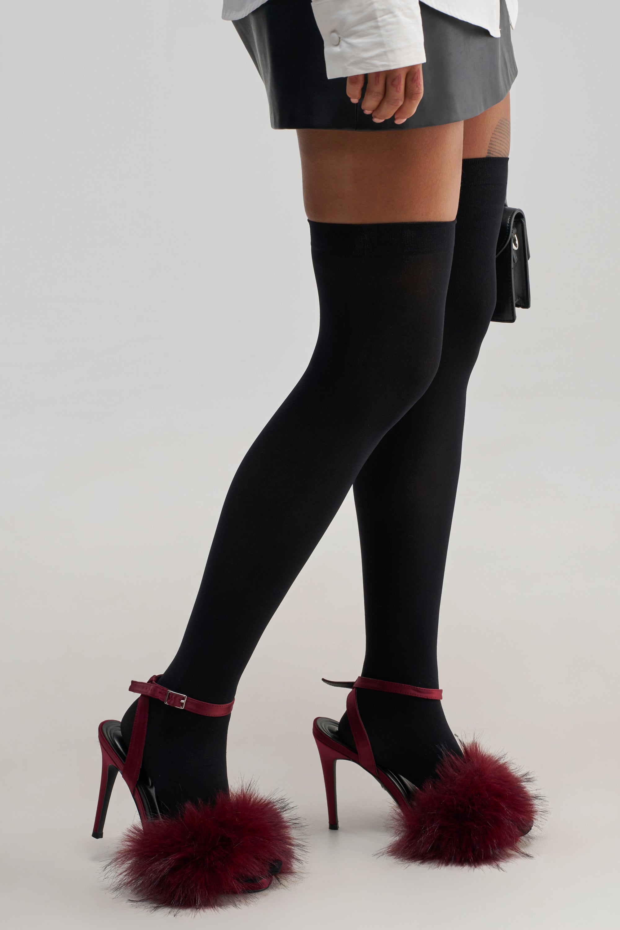 A woman styles the SEXY MESH THIGH-HIGH SOCKS with a black mini skirt, white shirt, and burgundy high heels featuring fluffy accents, creating an effortlessly chic fall layering look against a light background.