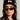 A person wearing the BOW PLEASE BEANIE, a black accessory embellished with satin bow appliques, and stylish sunglasses. They have long hair and are dressed in a denim jacket beneath a black jacket featuring an embroidered star. Their expression is thoughtful as they gaze slightly to the side.