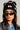 A person wearing the BOW PLEASE BEANIE, a black accessory embellished with satin bow appliques, and stylish sunglasses. They have long hair and are dressed in a denim jacket beneath a black jacket featuring an embroidered star. Their expression is thoughtful as they gaze slightly to the side.