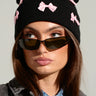 A person wearing the BOW PLEASE BEANIE, a black accessory embellished with satin bow appliques, and stylish sunglasses. They have long hair and are dressed in a denim jacket beneath a black jacket featuring an embroidered star. Their expression is thoughtful as they gaze slightly to the side.