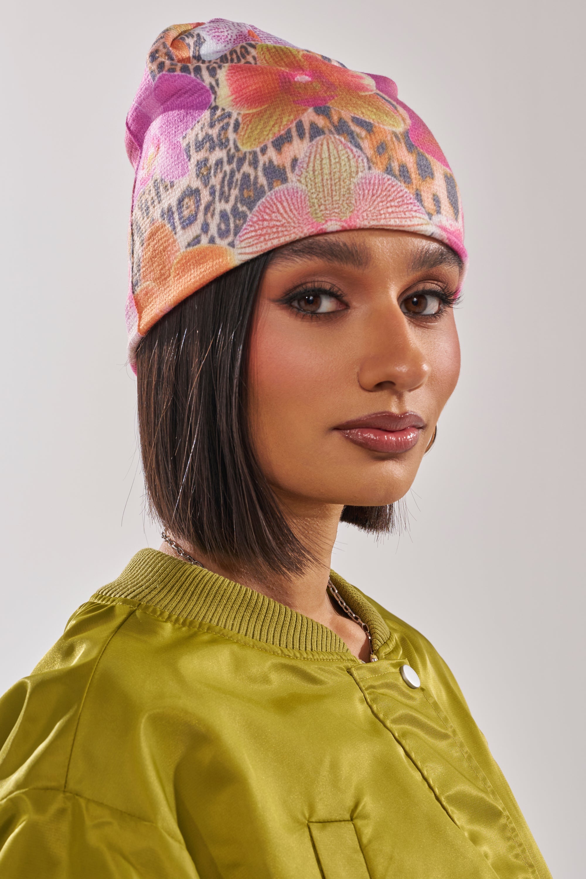 A woman with a sleek bob models street style, wearing the WILDLY FEMME LIGHTWEIGHT BEANIE in colorful floral leopard print and a shiny olive-green jacket, posing against a plain light background. 1