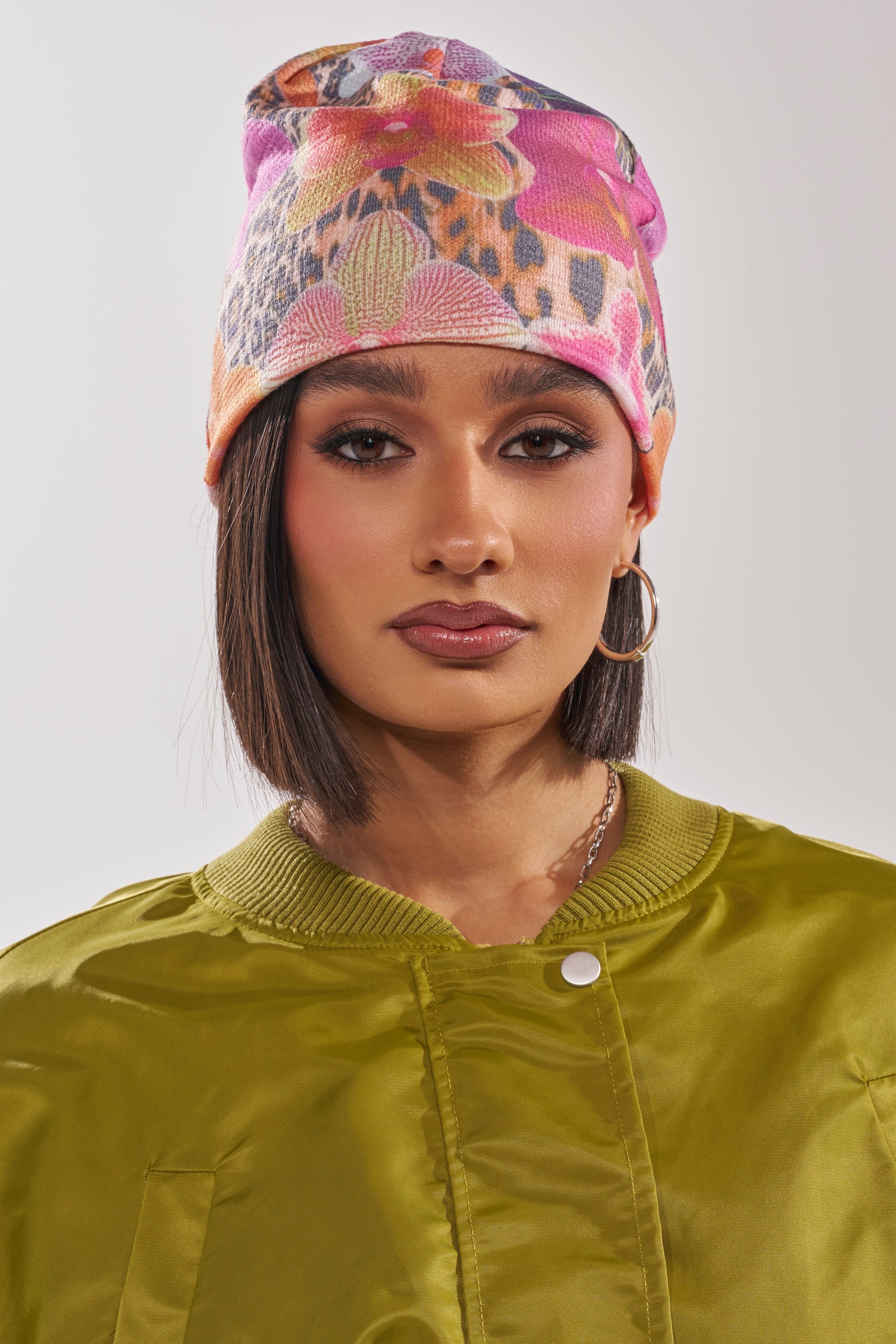 A woman with straight, shoulder-length dark hair wears the WILDLY FEMME LIGHTWEIGHT BEANIE featuring a trendy multicolor floral leopard print, along with gold hoop earrings and a shiny olive green jacket. She faces forward with a neutral expression. 2
