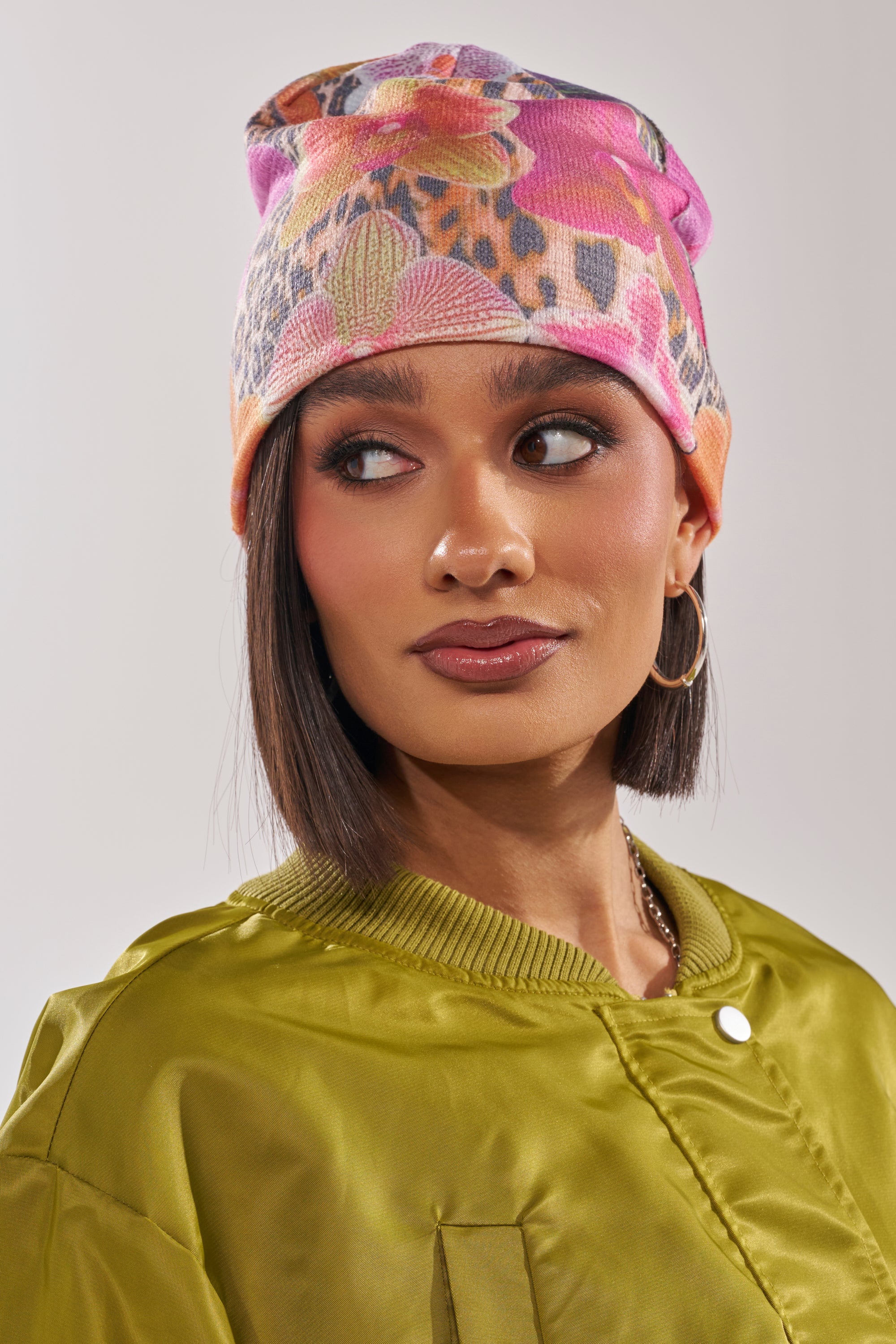 A woman with a straight brown bob models the WILDLY FEMME LIGHTWEIGHT BEANIE—a trendy multicolor floral leopard print style—paired with hoop earrings and a shiny olive green jacket, posing against a neutral background. 6