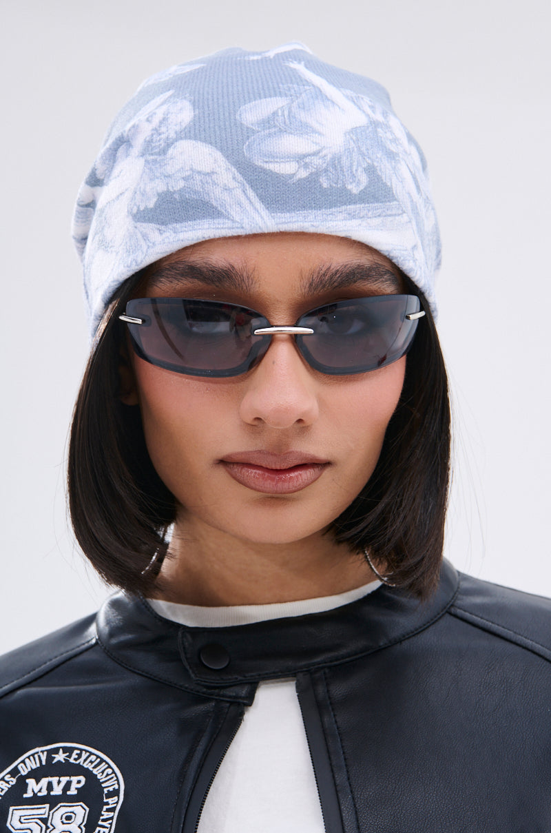 A person wears the CITY OF ANGELS PRINTED BEANIE—a light blue, patterned accessory—paired with dark rectangular sunglasses and a black leather jacket with a patch. They have straight, chin-length hair and a neutral expression. 2