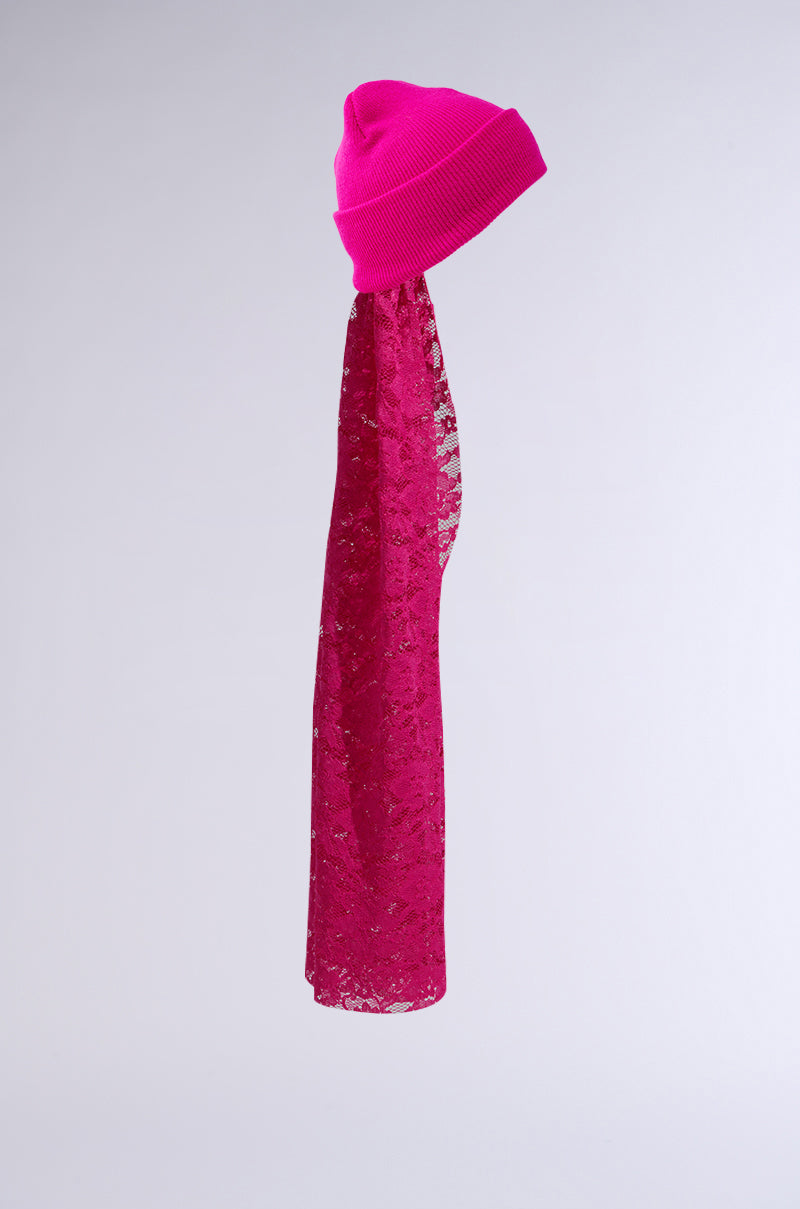 The IN THE CUT LACE VEIL BEANIE, a vibrant hot pink beanie featuring a long, sheer lace veil, hangs against a plain gray background. The veil drapes elegantly, showcasing its intricate lace design. 3