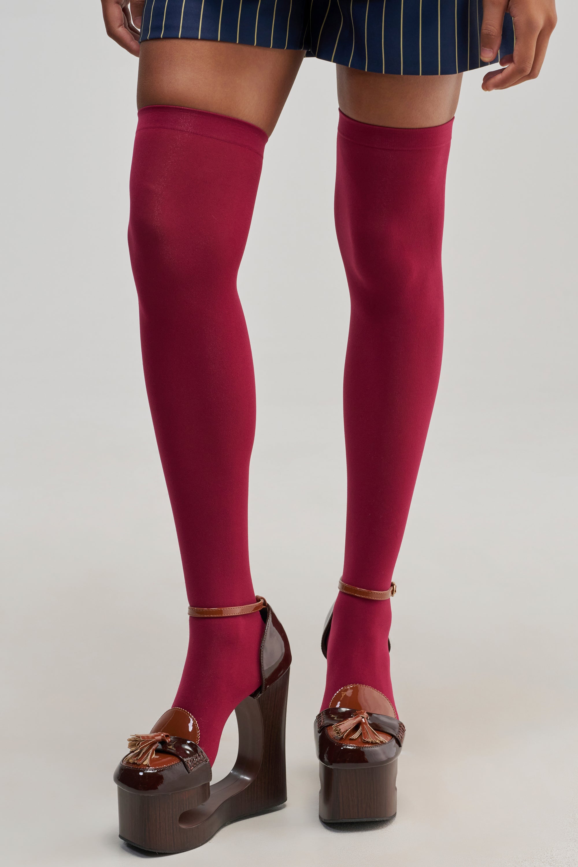 Wearing a romantic look, the person is styled in SHEER ATTITUDE SOCKS IN BURGUNDY with pinstriped navy shorts and brown platform heels featuring a glossy finish and decorative tassels, standing against a plain background.