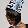 A person wearing the striking WILD ONES ZEBRA PATCH BEANIE, featuring a knit base with zebra patterns, blue patches, and silver cross embellishments. Their shoulder-length dark hair and thick silver chain necklace harmonize beautifully with their black top against a plain background.