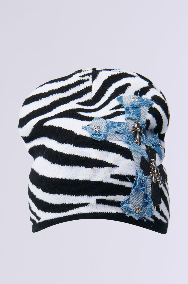 Introducing the WILD ONES ZEBRA PATCH BEANIE, this striking accessory showcases a black and white zebra design on a knit base. It is highlighted by a blue denim cross patch embellished with metal studs and small cross charms, all set against a neutral gray background. 2