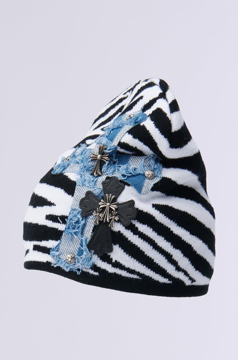 The WILD ONES ZEBRA PATCH BEANIE is a striking accessory featuring a knit base, adorned with zebra-patterned fabric and two cross-shaped patches—one crafted from distressed denim and another in black fabric with metallic accents—set against a light background. 3