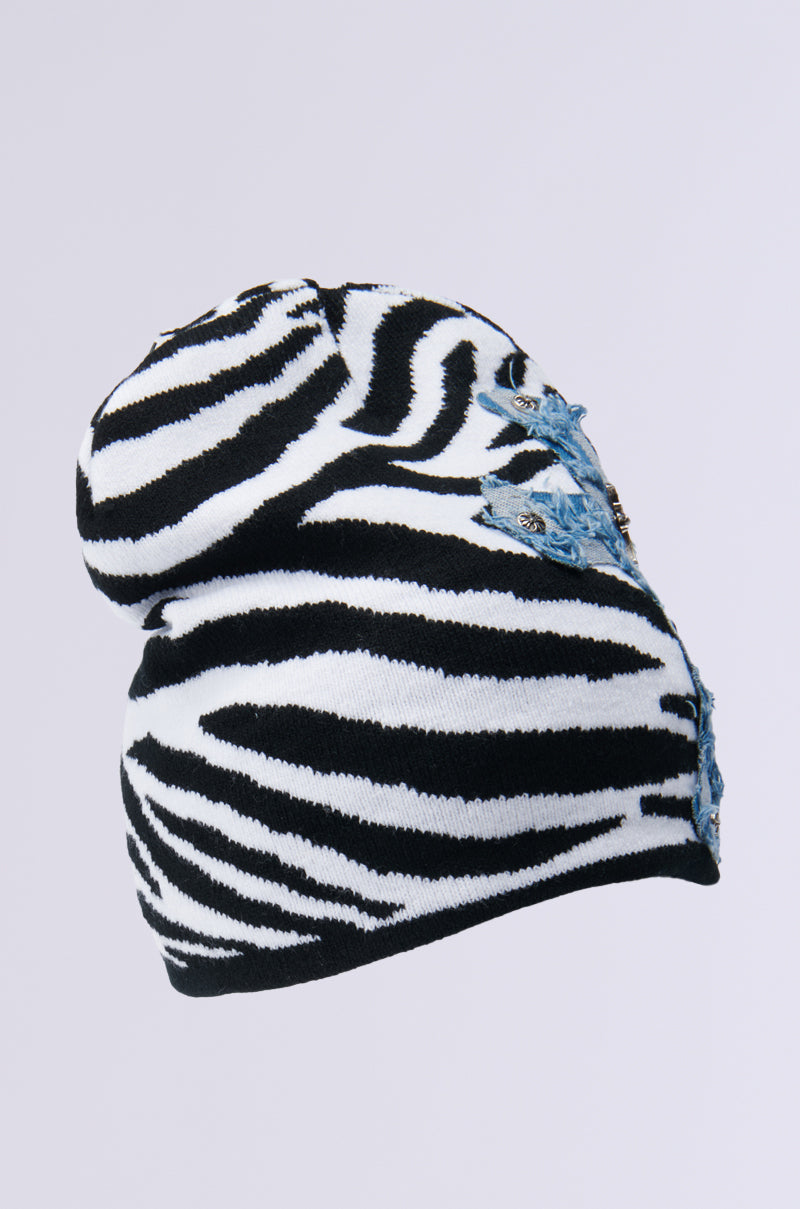 The WILD ONES ZEBRA PATCH BEANIE is a striking accessory featuring black and white zebra stripes on a knit base, embellished with blue floral embroidery and shimmering gems on the side, all set against a plain backdrop. 4