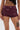 Wearing the HOT DAMN FAUX LEATHER SHORT WITH LACE UP SIDE in dark burgundy, this look features mid-rise faux leather shorts that highlight tattoos—“1990” on the abdomen and a face on the upper thigh—paired with a black lace crop top.