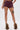 Wearing HOT DAMN FAUX LEATHER SHORT WITH LACE UP SIDE in burgundy, styled with beige furry boots and a skull thigh tattoo, the person poses against a plain white background.