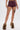Someone wears the HOT DAMN FAUX LEATHER SHORT WITH LACE UP SIDE in burgundy with a mid-rise fit, paired with tan high-heeled shoes trimmed in brown faux fur. A skull tattoo appears on their upper left thigh against a plain white background.