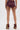 A person stands with their back to the camera, wearing HOT DAMN FAUX LEATHER SHORT WITH LACE UP SIDE in burgundy, paired with matching fur-trimmed high-heeled boots and showcasing ballet shoe tattoos on the backs of their thighs.