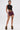 A person stands confidently in a black lace crop top, HOT DAMN FAUX LEATHER SHORT WITH LACE UP SIDE, furry boots, and shows tattoos on their torso and thigh against a plain white background.