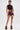 A woman stands confidently in HOT DAMN FAUX LEATHER SHORT WITH LACE UP SIDE, paired with a black lace crop top and fluffy brown ankle boots. She shows off bold tattoos and large hoop earrings, posing against a plain white background.