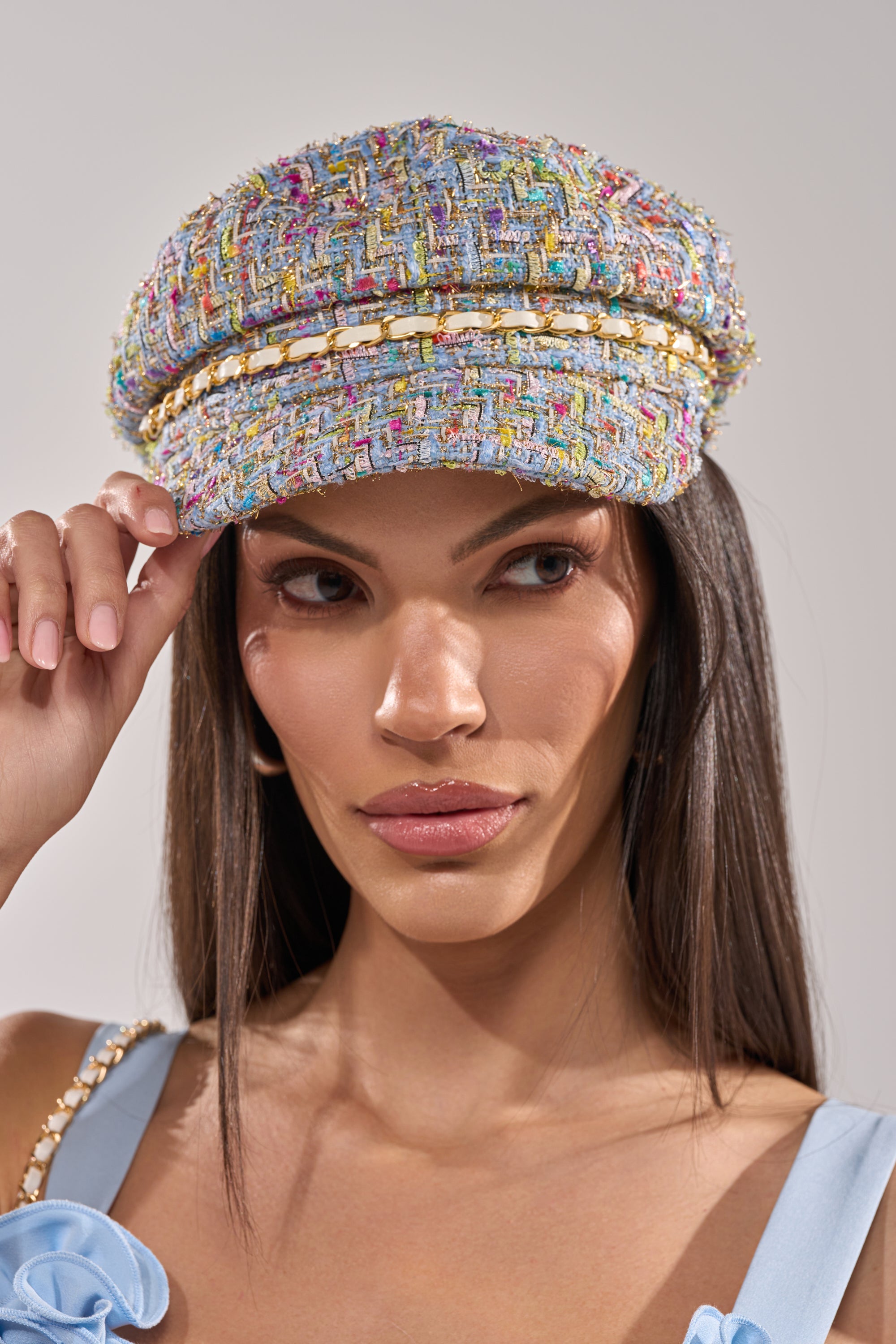 A woman with long dark hair wears the I HAVE CLASS TWEED BRIM HAT, a chic multicolor newsboy cap with gold chain trim, paired with a light blue top featuring a bow strap, as she looks to the side and adjusts her hat. 0