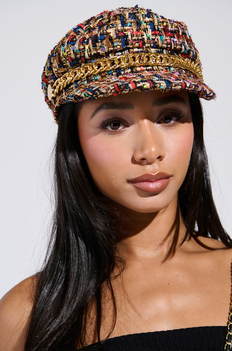 A woman with long dark hair wears the IN THE CITY BRIM HAT, styled like a baker boy cap with gold accents, paired with a black strapless top. She's set against a white background, looking calm and composed.
