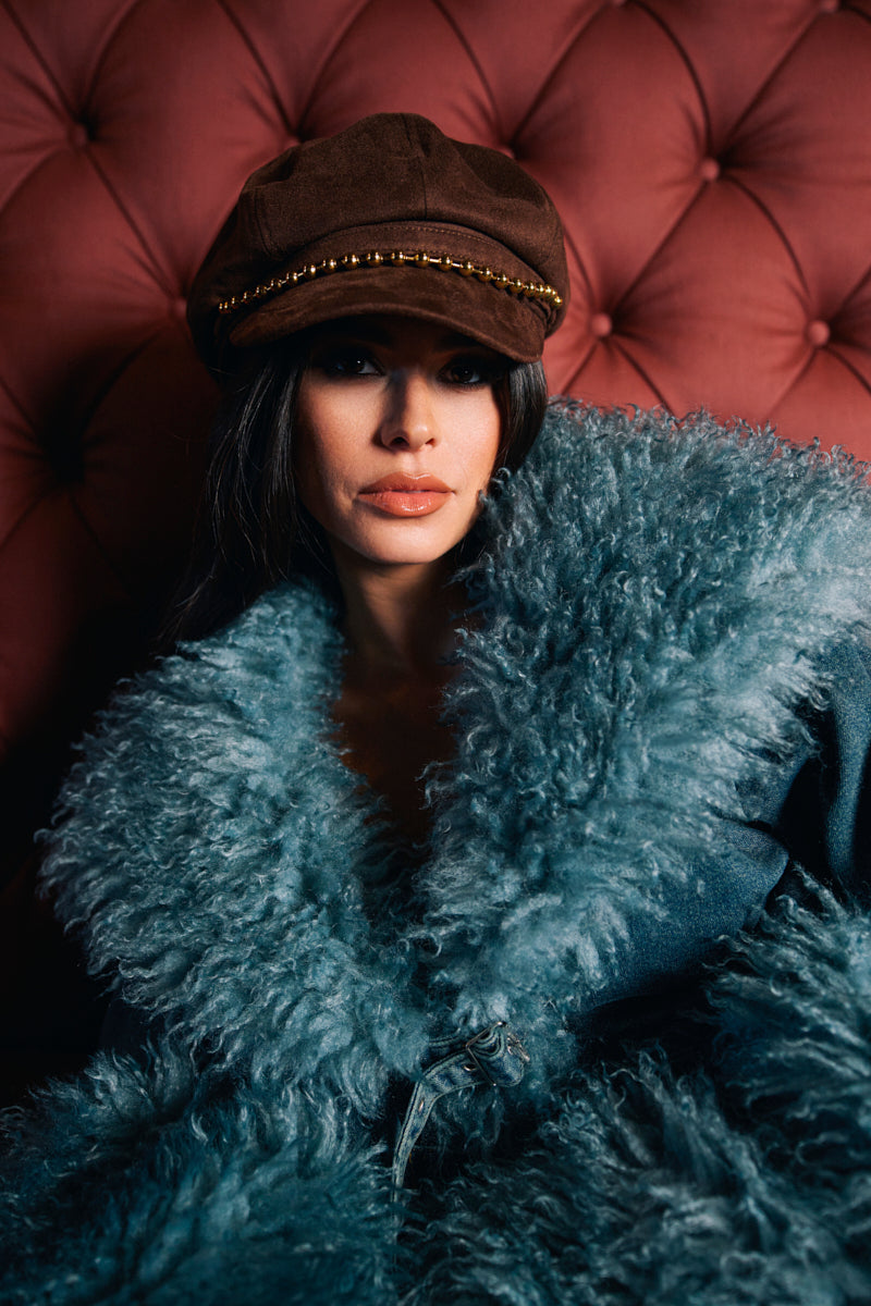 A woman wearing the IT GURL BRIM HAT and a teal faux fur coat sits against a tufted maroon background, looking confidently at the camera. 0