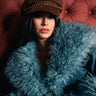 A woman wearing the IT GURL BRIM HAT and a teal faux fur coat sits against a tufted maroon background, looking confidently at the camera.