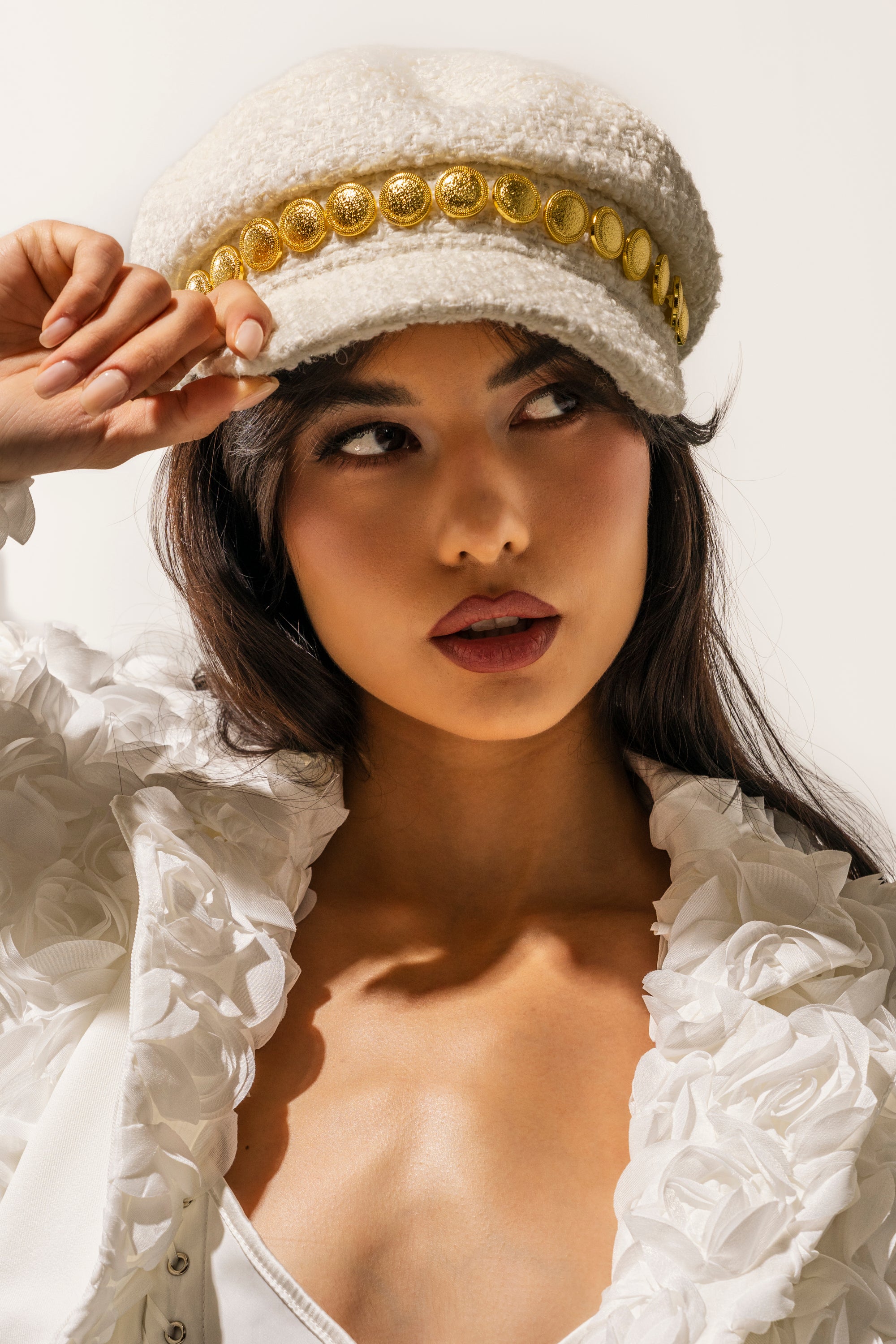 A woman in a white ruffled top holds the brim of the IN MALLORCA TWEED BRIM HAT, an ivory newsboy cap detailed with gold buttons, as she gazes to the side against a light background. 0