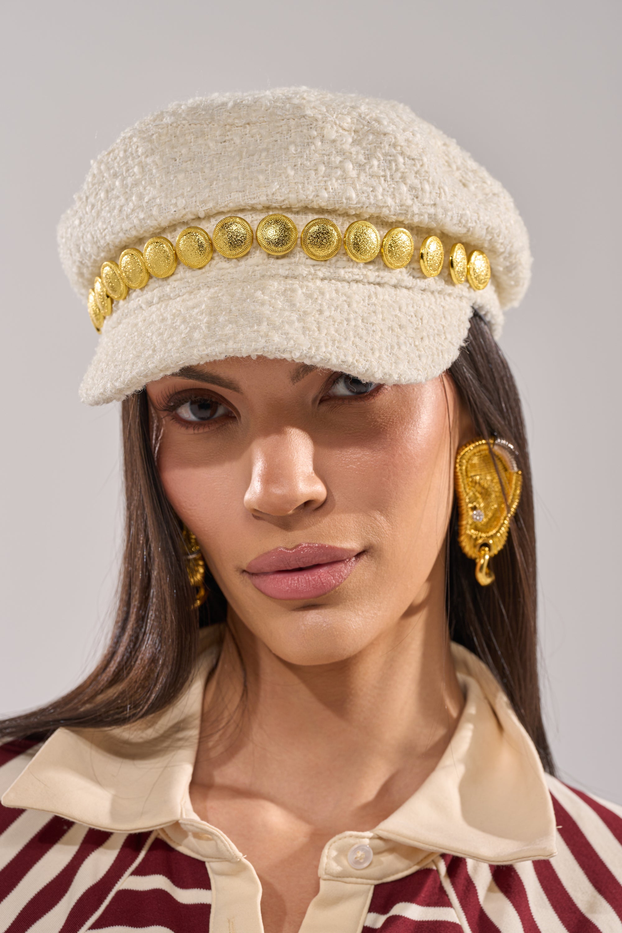 A woman with long brown hair and a neutral expression wears the IN MALLORCA TWEED BRIM HAT, gold statement earrings, and a beige and maroon collared shirt with a geometric pattern. 1