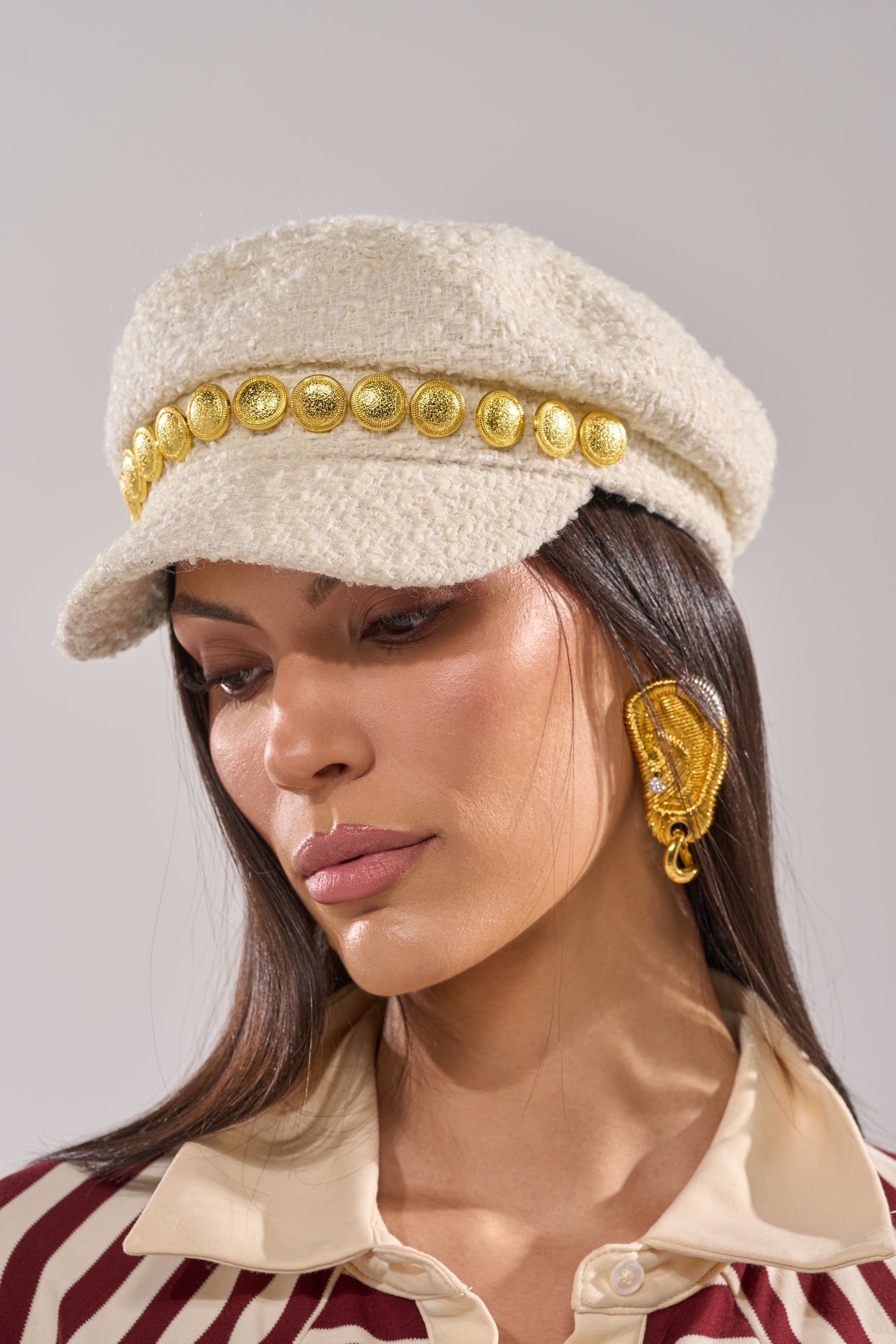 A woman wears the IN MALLORCA TWEED BRIM HAT in ivory, styled with gold leaf earrings and a maroon-and-beige collared shirt, looking downward against a neutral background. 2