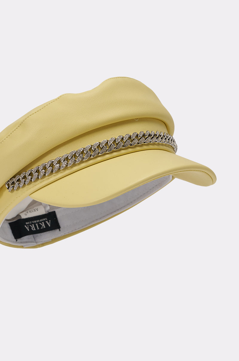 The BUTTER ME UP PU BRIM HAT is a butter yellow faux leather newsboy cap with a curved visor and sparkling rhinestone chain trim above the brim. The interior label reads "AKIRA. 6