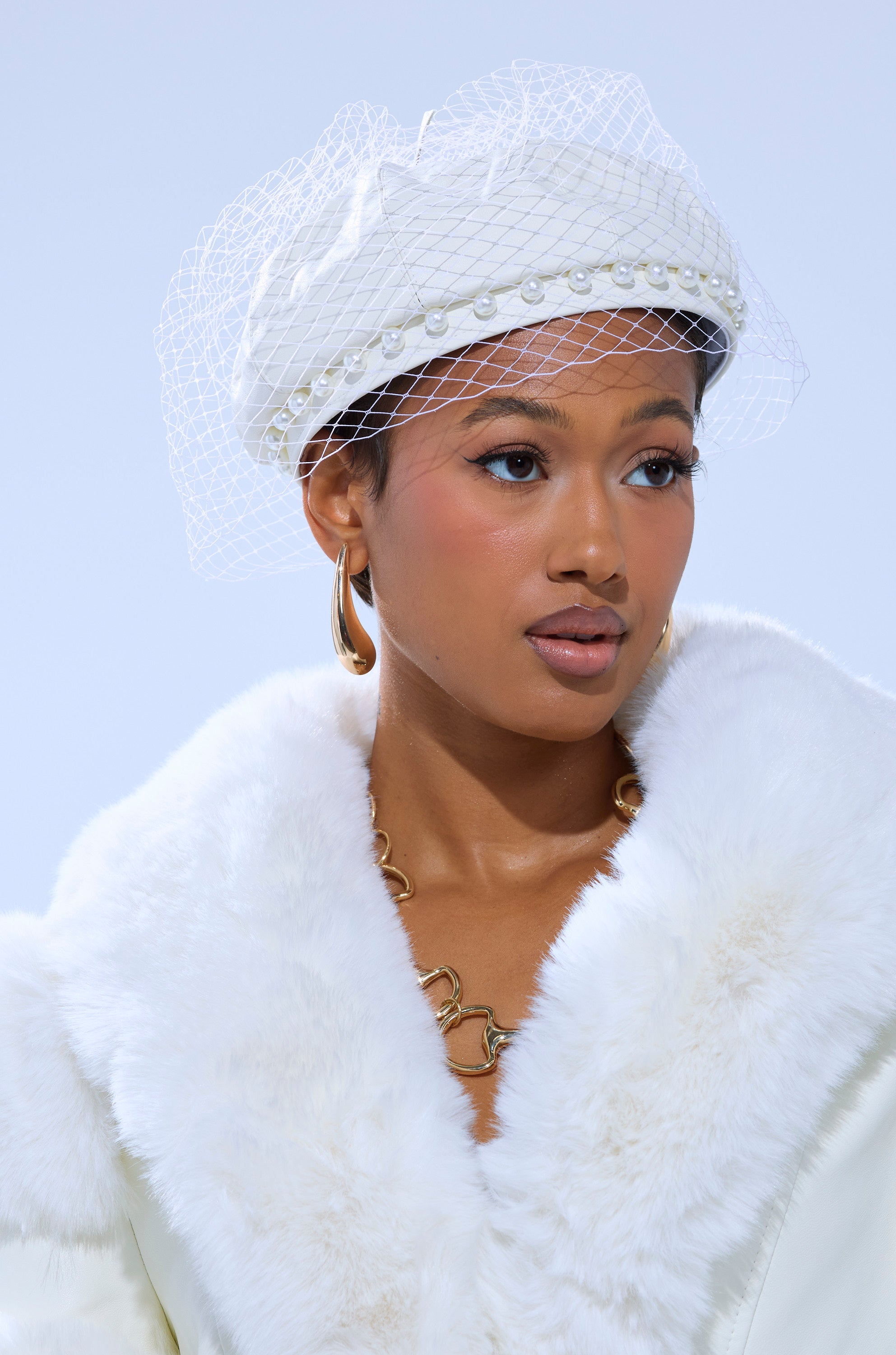 A woman wears a white fur coat and the EDITORIAL BERET featuring pearls and a mesh veil, styled with gold hoop earrings, a gold chain necklace, natural makeup, pink blush, and set against a light blue background.