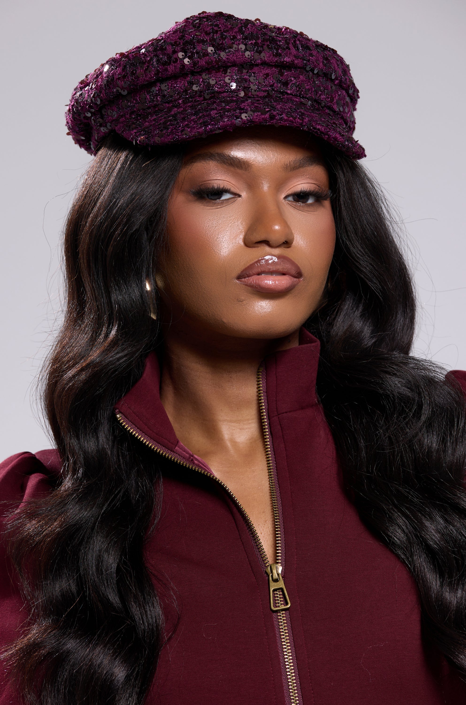 A woman with long, wavy dark hair wears the KARY TWEED HAT in purple with a matching zip-up top. Her makeup is polished, featuring glossy lips and defined brows, as she gazes confidently at the camera against a plain background. 1