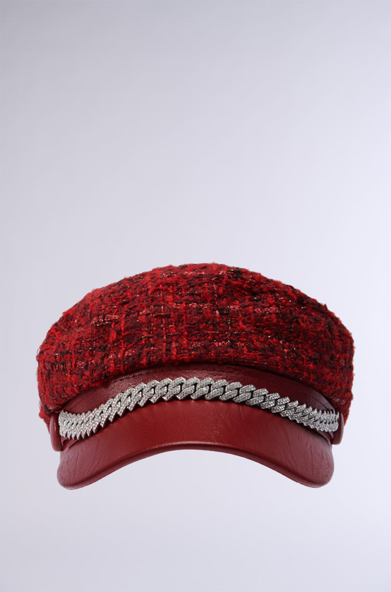 The Tweedy Baker Boy Cap in Red, adorned with metallic accents and a silver chain detail above the brim, stands out against a plain gray background. 2