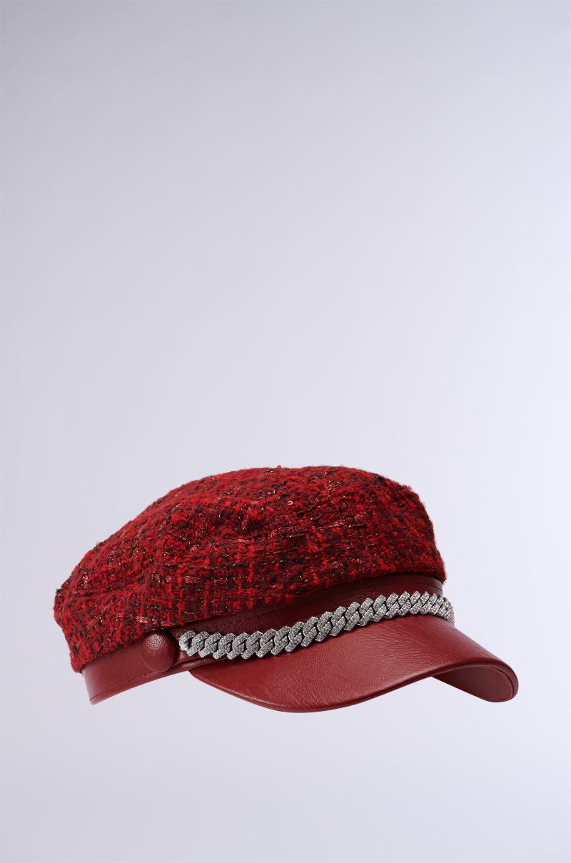 The TWEEDY BAKER BOY CAP IN RED is a chic statement piece, showcasing a stylish tweed design with a leather brim and a decorative silver chain featuring metallic accents. Positioned against a light gray background, this cap enhances any wardrobe with its fashionable appeal. 3